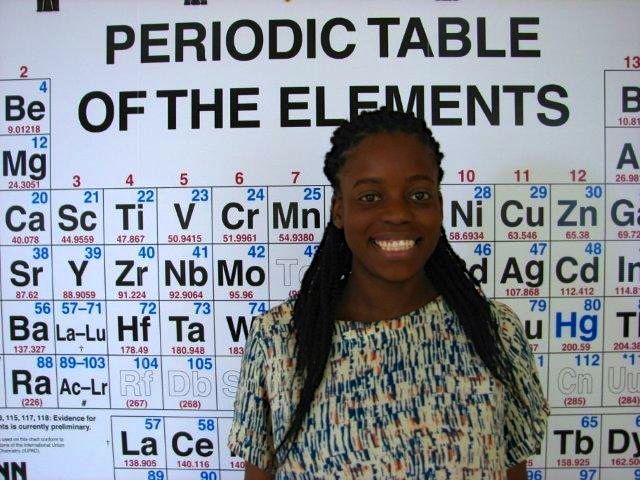 ​Mrs. Miller poses beside her classroom copy of the Periodic Table.